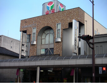株式会社山元百貨店（当時）の外観。手前にアーケードがあり、「YAMAMOTO」や「山元」という多数の表記で看板が掲げられている